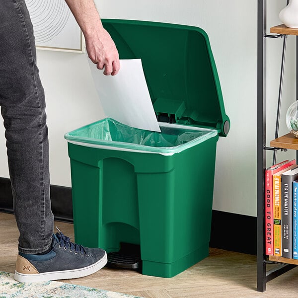 A person standing next to a Lavex green rectangular step-on trash can and putting paper in it.