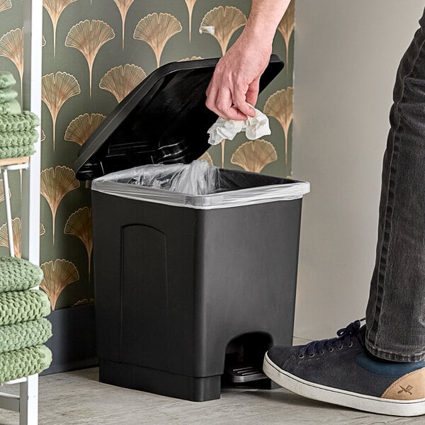 A person throwing a tissue into a Lavex black rectangular step-on trash can.
