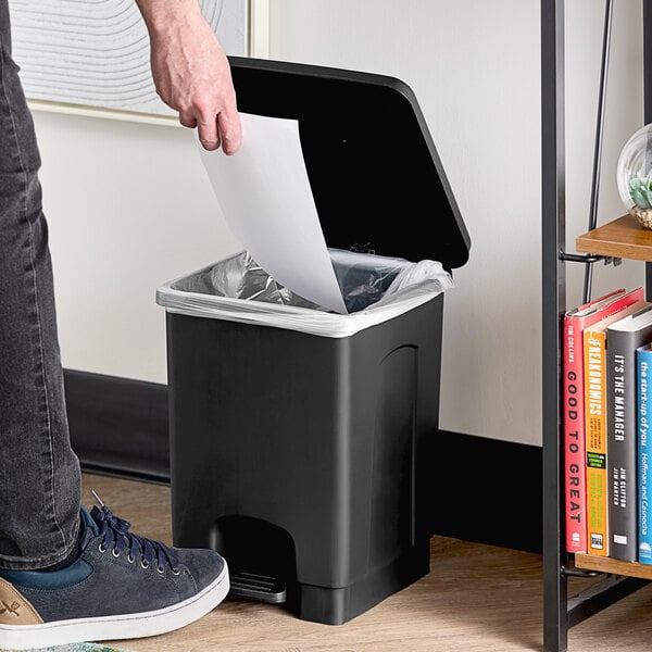 A person standing next to a Lavex black rectangular step-on trash can with a white paper in hand opening the lid to throw it away.