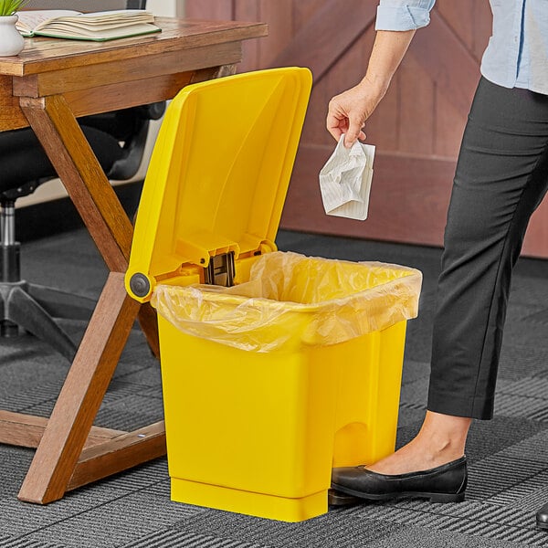 A woman's hand throws paper into a yellow Lavex step-on trash can next to a wooden table.