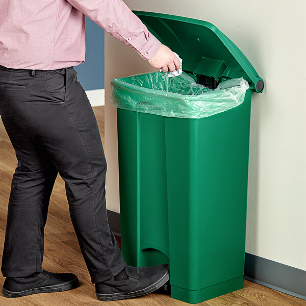 A man in black pants standing next to a Lavex green rectangular step-on trash can with a plastic bag inside.