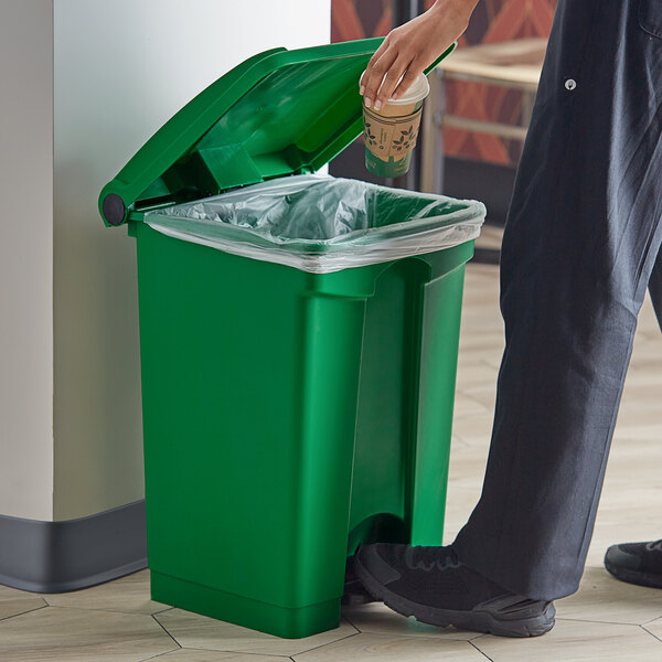 A person using a hand to open a green Lavex step-on trash can lid to put a coffee cup in it.