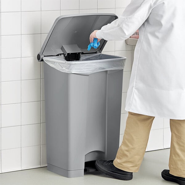 A person in a lab coat and gloves opening a Lavex rectangular step-on trash can and putting a blue glove inside.