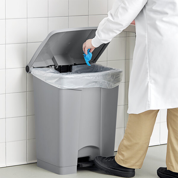 A person in a white coat putting a blue glove in a Lavex step-on trash can.