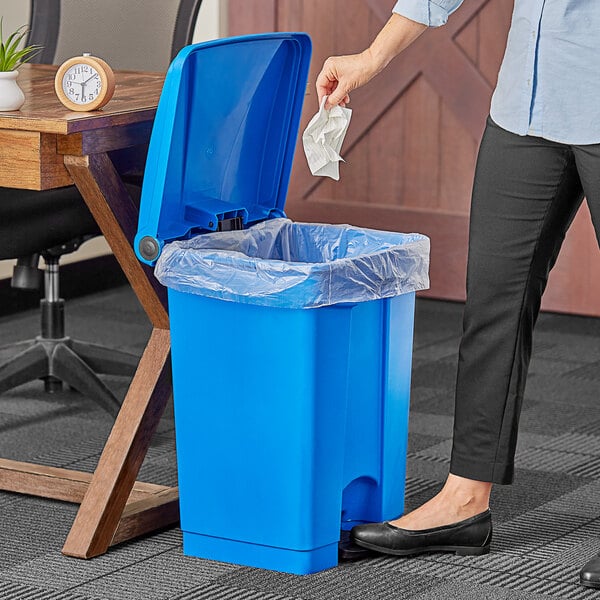 A woman using a Lavex blue rectangular step-on trash can to throw paper in.