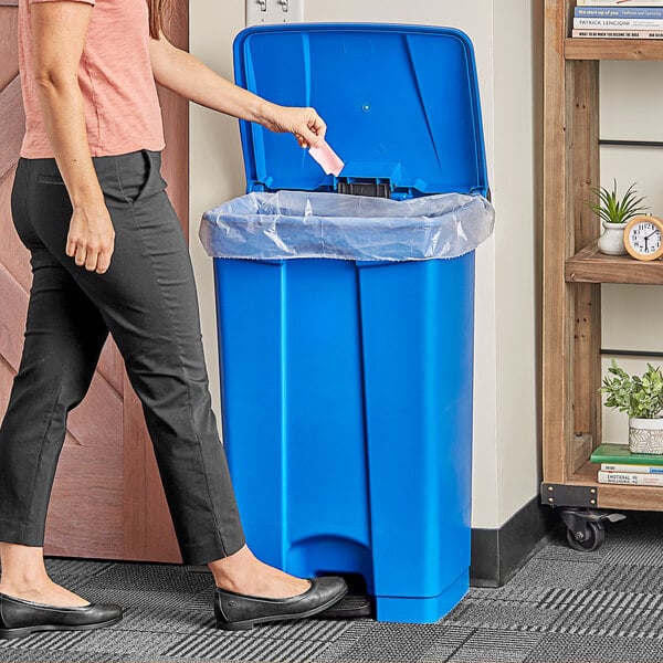 A woman using a Lavex blue rectangular step-on trash can to throw away paper.