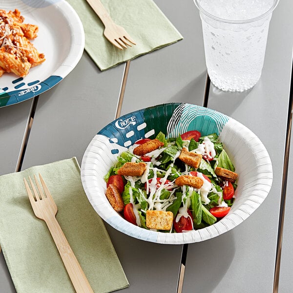 A bowl of salad with croutons and tomatoes in a Choice heavy weight paper bowl on a table with a wooden fork and clear plastic cup of water.