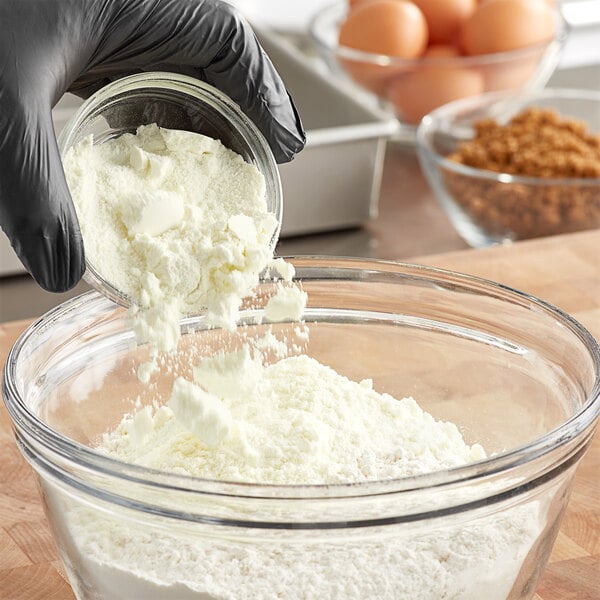 A gloved hand pouring Judee's Whole Milk Powder into a bowl of flour.