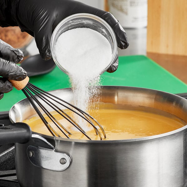 A hand in gloves pouring Judee's From Scratch Sodium Citrate Powder into a pot of liquid.