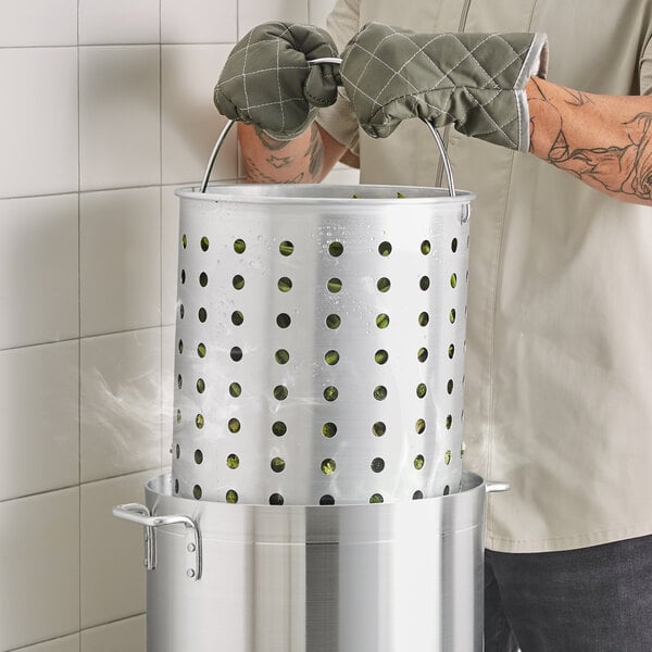 A man using gloves to hold a Choice aluminum stock pot with food in a steamer basket.