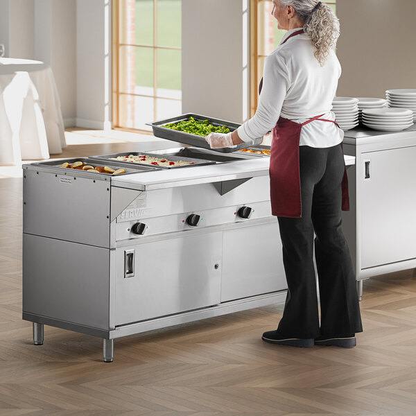 A woman wearing a red apron and holding a tray of food standing in front of a ServIt Electric Steam Table.