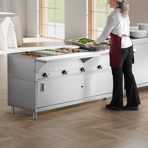 A woman in a red apron preparing food in a commercial kitchen using a ServIt electric steam table.