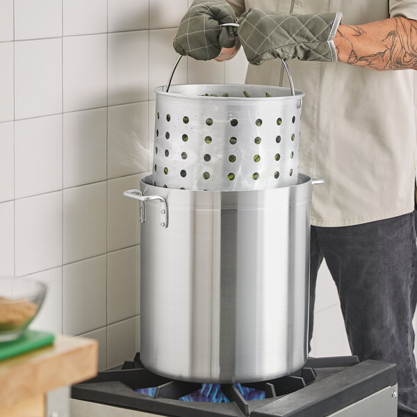 A man using gloves to hold a Choice aluminum stock pot with a steamer basket over a stove.