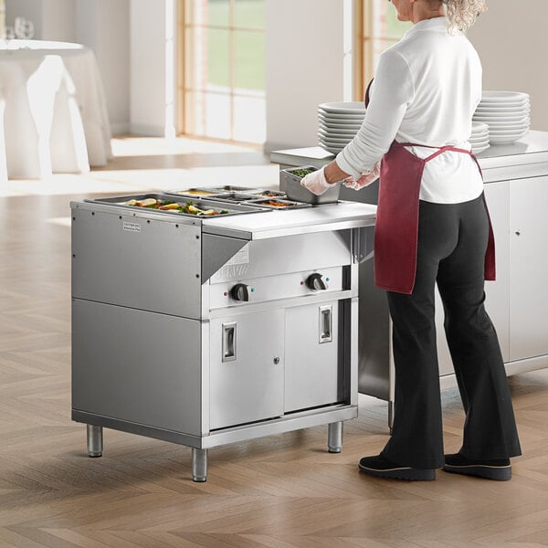 A woman wearing a red apron cooking food in a commercial kitchen using a ServIt electric steam table.