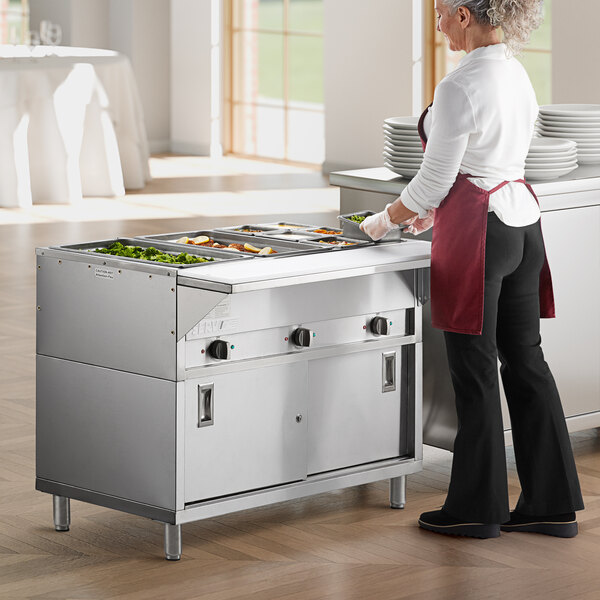 A woman in a red apron and gloves using a ServIt electric steam table in a commercial kitchen.