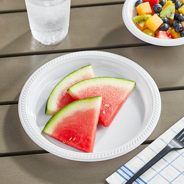 A white plastic plate with watermelon slices and fruit on a table with a fork.