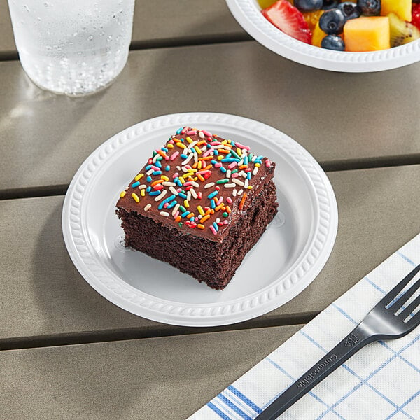 A Choice white plastic plate with chocolate cake and fruit on it, next to a fork and a glass of water.