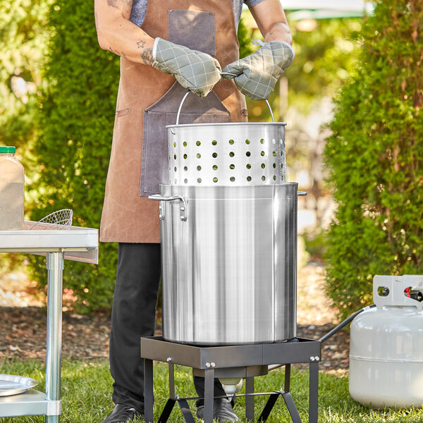 A man in an apron and gloves using a Choice aluminum stock pot with a steamer basket and cover to cook outdoors.