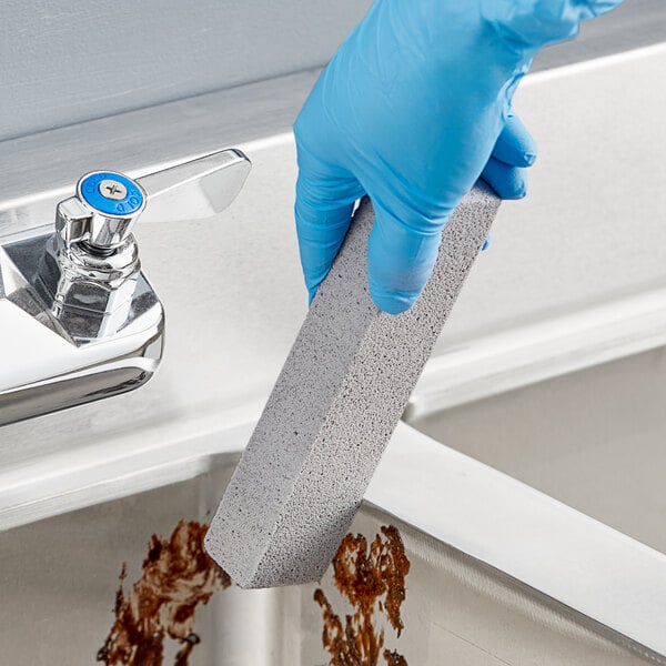 A person wearing blue gloves using a Lavex Pumice Stone to clean a sink.