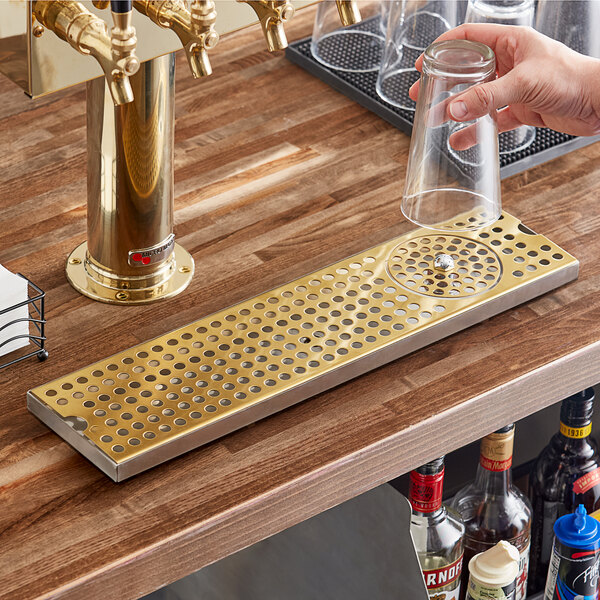 A hand holding a glass over a metal bar with a Regency PVD gold finish surface-mount beer drip tray.