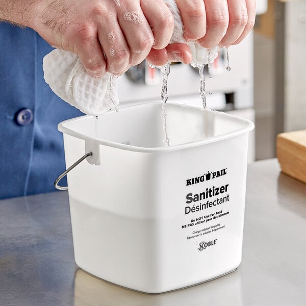 A person washing dishes in a white Noble Products sanitizing pail on a counter.