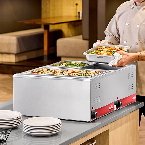 A man in a chef uniform using an Avantco countertop food warmer to hold food in containers on a table.