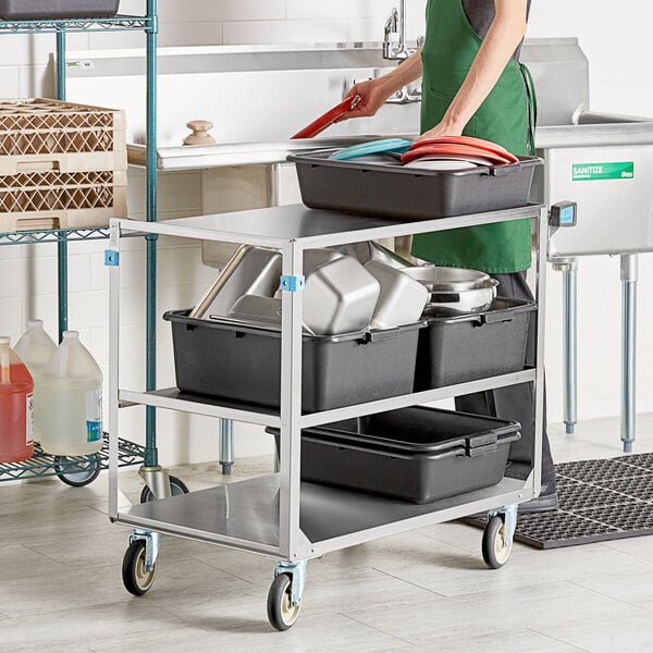 A woman in a green apron using a Lakeside stainless steel utility cart to transport black, grey, and red containers.