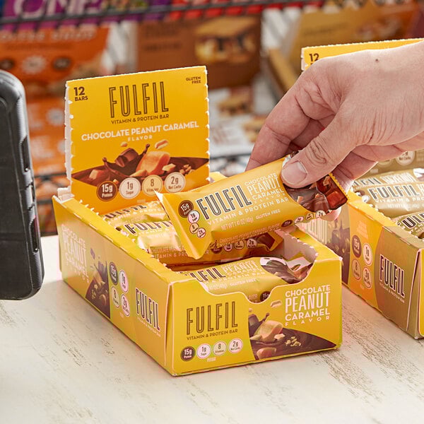 A hand holding a box of FULFIL Chocolate Peanut Caramel Vitamin and Protein Bars.