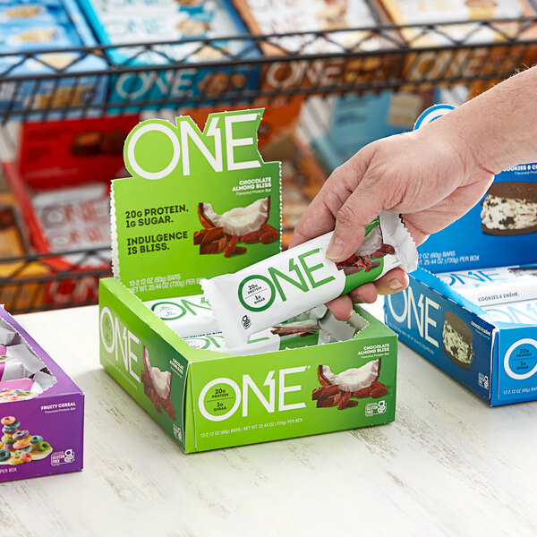 A person putting a Chocolate Almond Bliss ONE Protein Bar in a green box with white text.
