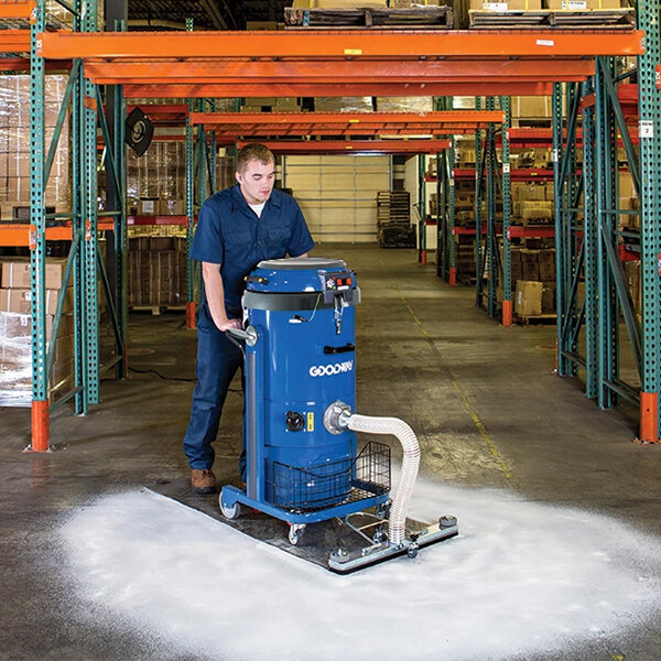 A man in blue overalls using a blue Goodway walk-behind dry vacuum on a warehouse floor.