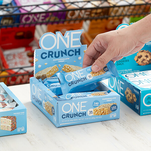 A hand holding a blue and white box of ONE Crunch Marshmallow Treat protein bars.