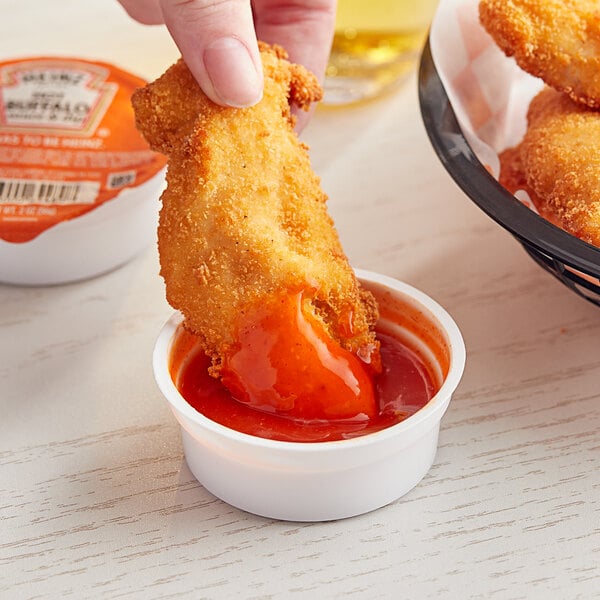 A person dipping a chicken wing into a small container of Heinz Hot Buffalo Sauce.