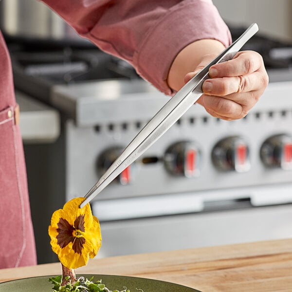 A person using Mercer Culinary plating tongs to place a knife on a plate of food.