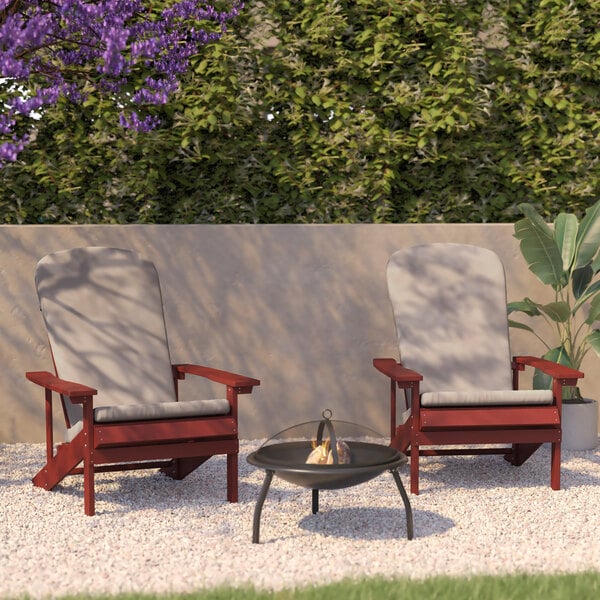 Two red Flash Furniture Adirondack chairs with cream cushions next to a black metal fire pit on a gravel path.