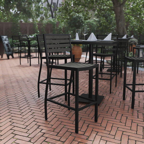A Flash Furniture Lark Gray Wash faux teak slat stackable barstool on a brick patio with a table and wine glasses.