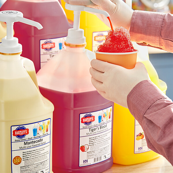 A hand holding a cup of red liquid with a container of Hartley's Tiger's Blood Snow Cone Syrup.