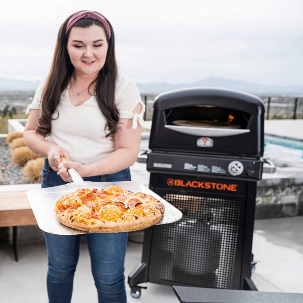 A woman holding a pizza in front of a Blackstone outdoor pizza oven on a stand.