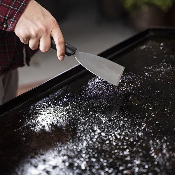 A hand using a Blackstone Griddle cleaning tool to scrape a black surface.