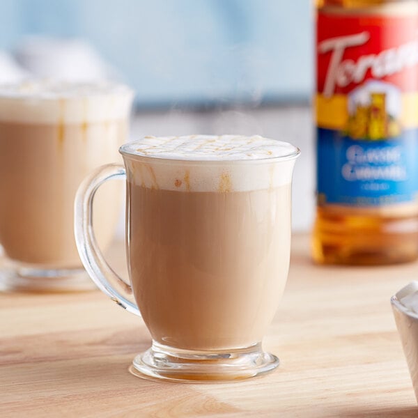 A glass mug of coffee with Torani Classic Caramel Syrup next to a bottle of it.