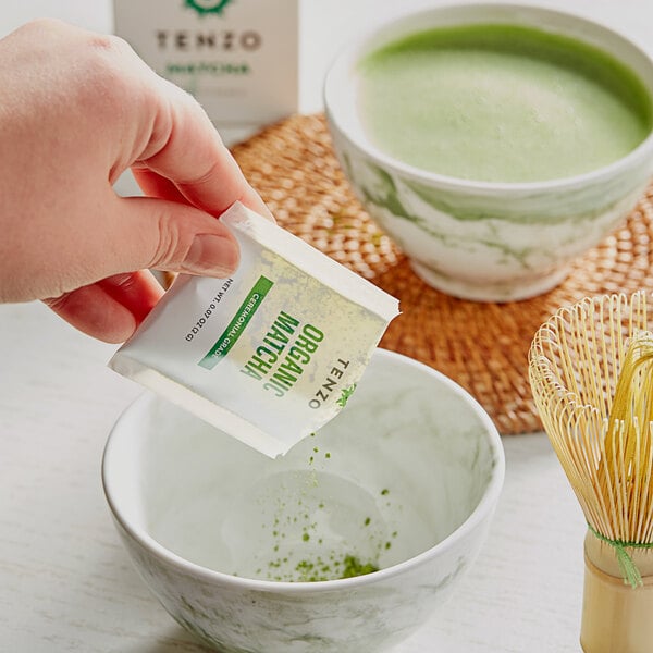 A hand pouring Tenzo Organic Ceremonial Matcha green tea powder from a white packet into a bowl.