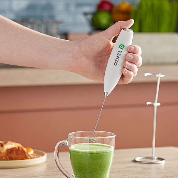 A person using a white Tenzo handheld milk frother to mix a green drink in a glass.