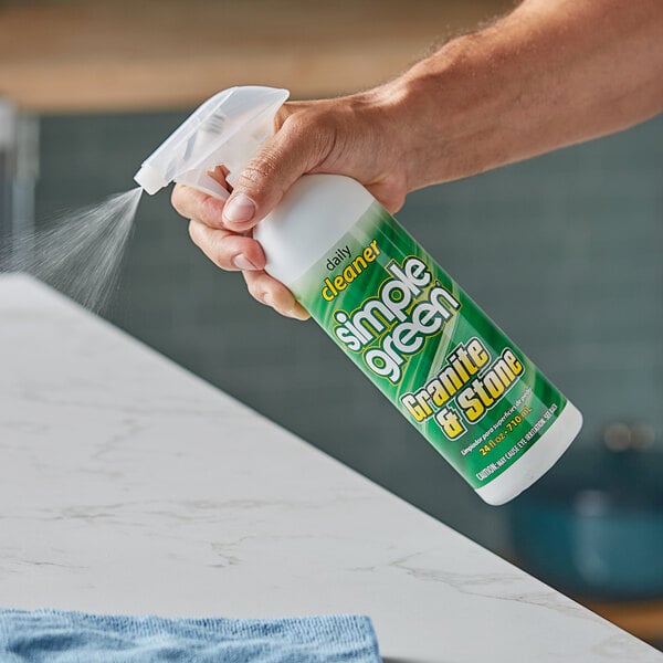 A hand holding a green and white Simple Green spray bottle spraying cleaner on a granite counter.