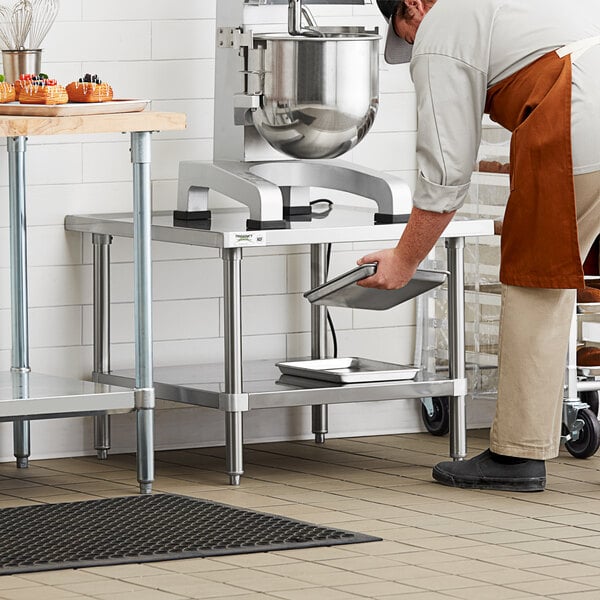 A man in a kitchen standing next to a Regency stainless steel mixer table with a metal container of whisks.