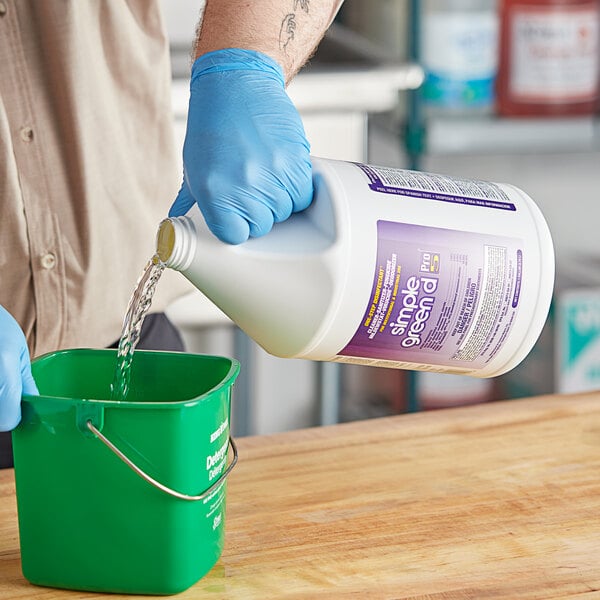 A person in blue gloves pouring Simple Green D Pro 5 disinfectant liquid into a green bucket.
