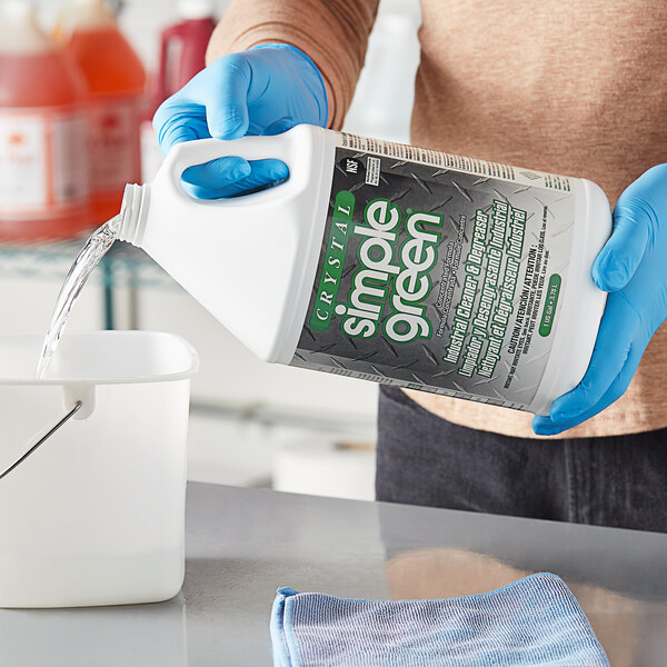 A person in blue gloves pouring Simple Green Crystal industrial cleaner from a jug into a white container.