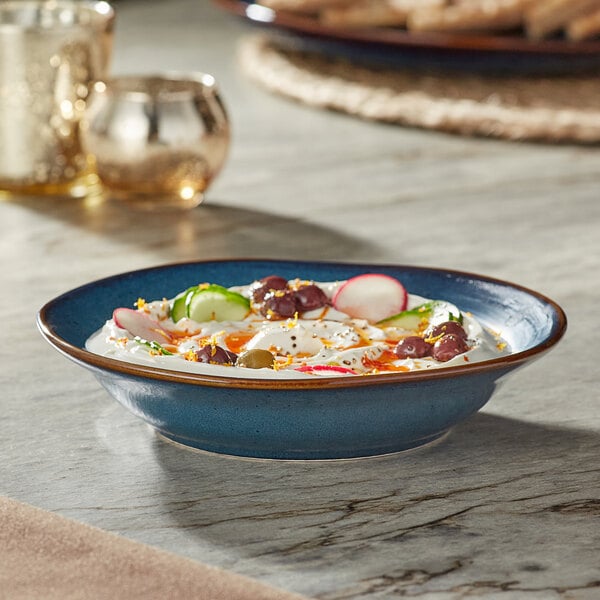 An Acopa Highlands skyfall blue china bowl filled with hummus surrounded by crackers and vegetables on a table.