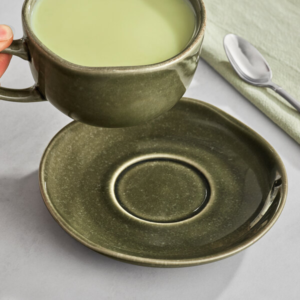 A hand holding a cup of green tea over a juniper green Acopa Highlands saucer on a table.