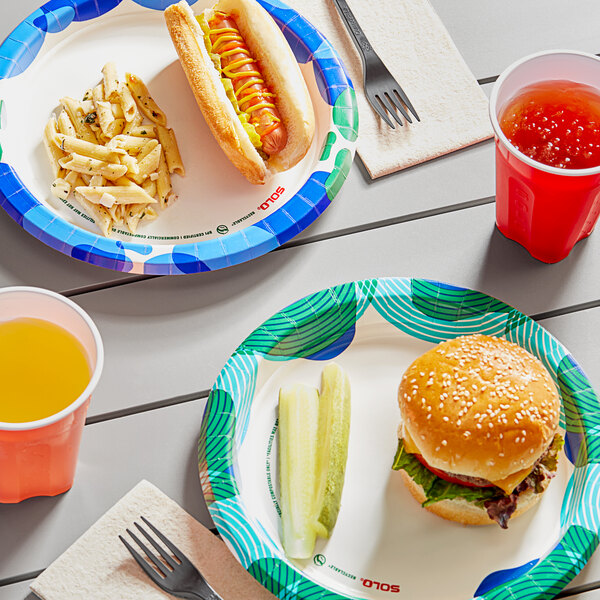 A Solo paper plate with a hamburger, hot dog, and a cup of orange liquid on a table.