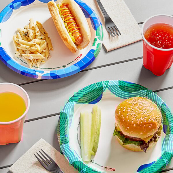 A Solo paper plate with a hamburger, hot dog, and cup of orange liquid on a table.