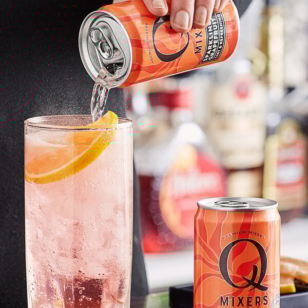 A hand pours Q Mixers Sparkling Grapefruit from a can into a glass with a slice of orange.